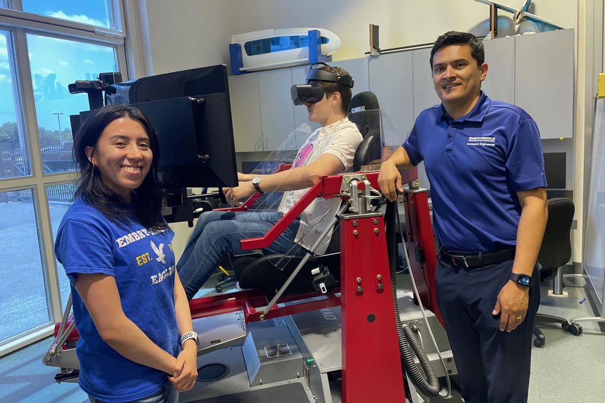 Embry-Riddle researcher Dr. Hever Moncayo (right) and graduate student Gabriela Gavilánez Gallardo (left) stand in the Advanced Dynamics and Control Lab next to the flight simulator being used for their AI research project. Aerospace engineering Ph.D. student Nathan Schaff, who is also a pilot, is sitting in the flight simulator.
