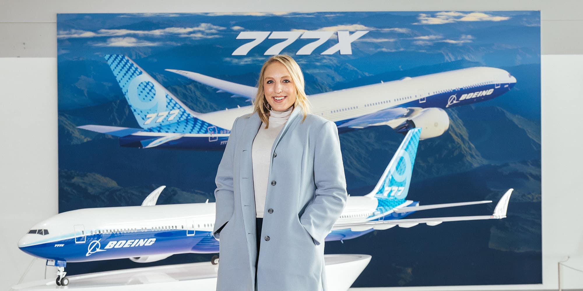 Rachelle Strong, wearing a light blue trench coat and a white turtleneck, stands in front of a backdrop showing several Boeing 777Xs flying over mountains.