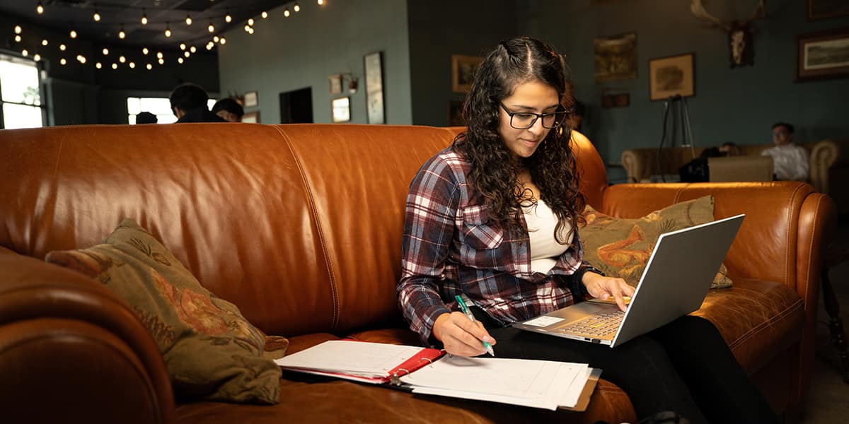 A student sits with a laptop open on her lap, sitting in an overstuffed leather sofa.